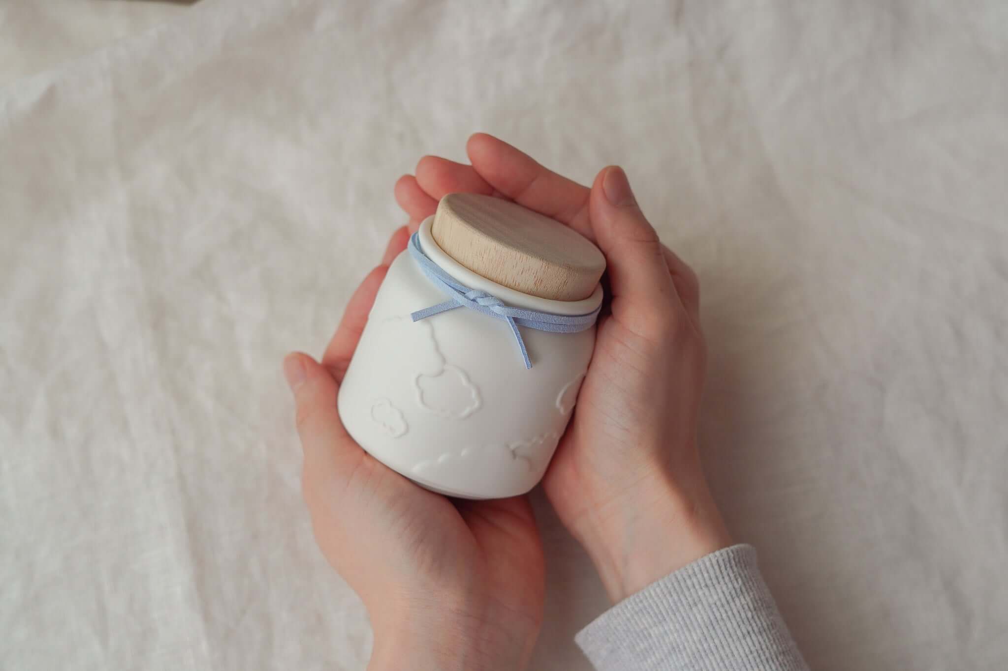 赤ちゃんのお骨壷を手に持つ優しい手の画像。白い陶器に木の蓋、雲の模様が施されています。